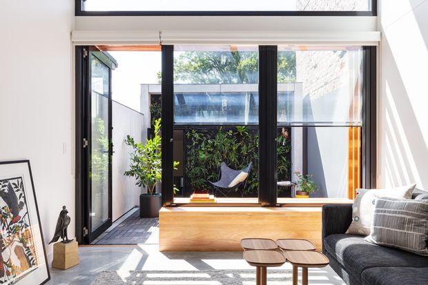 Bright contemporary living room with a large black-framed glazed opening onto a compact courtyard. The room features a built-in light timber window seat or storage bench, dark upholstered sofa, nesting timber coffee tables, indoor plants, framed artwork, and strong sunlight creating geometric shadows across the pale floor. A glazed courtyard structure and garden are visible beyond.