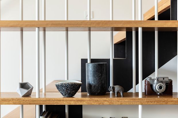 Close-up interior detail of open timber shelving with slender white vertical metal rods, displaying sculptural ceramics, a small animal figurine, and a vintage camera. A black geometric stair or wall element is visible behind the shelving, creating a graphic, modern composition.