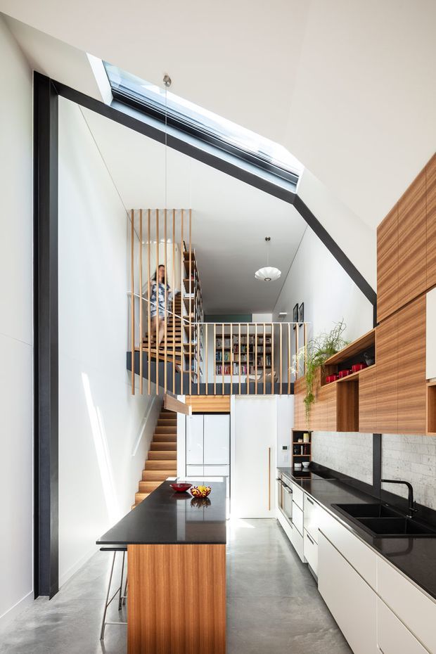 A bright, narrow contemporary kitchen beneath a dramatic sloping ceiling, with a long black island, white lower cabinetry, warm timber overhead joinery, dark benchtops, and a pale stone or tiled splashback. The kitchen opens visually toward a timber staircase and mezzanine library area with slim vertical balustrades. A large linear skylight brings daylight into the double-height space, while strong black framing and exposed concrete flooring add contrast.