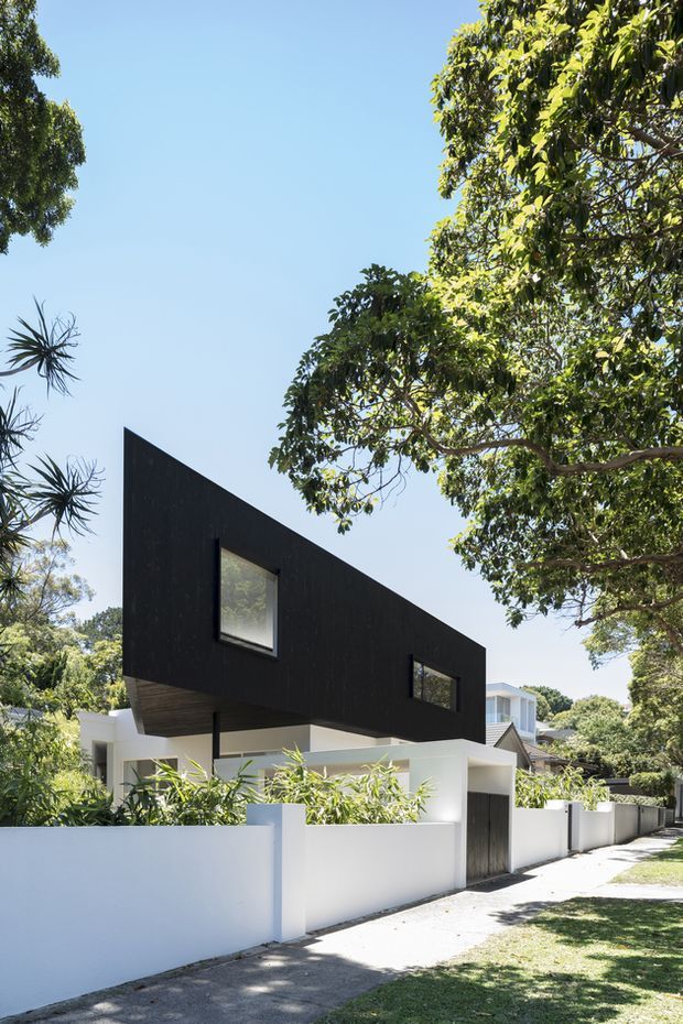 Street-facing exterior photograph of a contemporary two-storey residence in a leafy suburban setting. The house features a dramatic angular black upper volume with a sharply sloping roofline, narrow dark-framed windows, white rendered lower walls, a white boundary wall, and a recessed dark timber-toned entry gate. Mature trees and tropical planting frame the property.