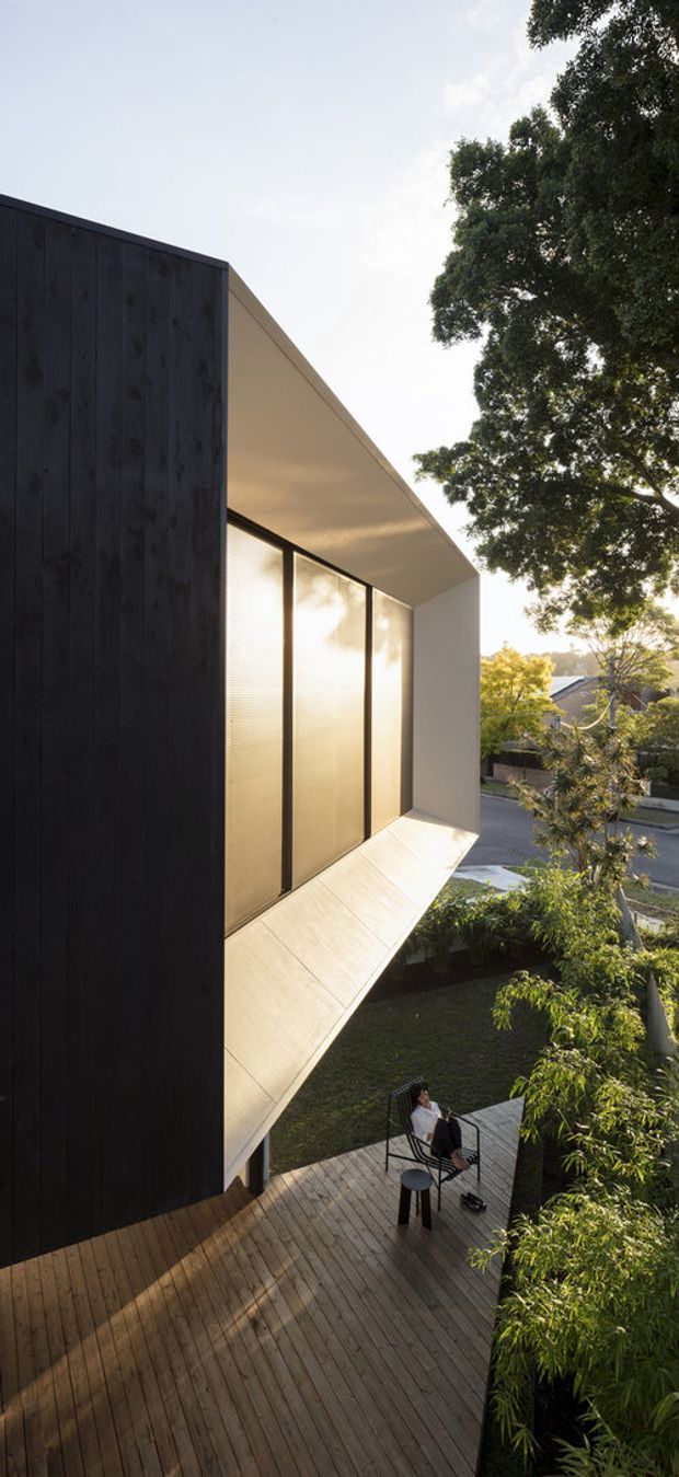 A vertical exterior architectural photograph showing a contemporary two-storey residence with a dramatic cantilevered upper volume. The facade combines dark vertical timber cladding with large translucent or reflective glazed panels framed in dark metal, beneath a clean white soffit. A timber deck and landscaped garden sit below, with outdoor seating and mature trees surrounding the home in a suburban setting.