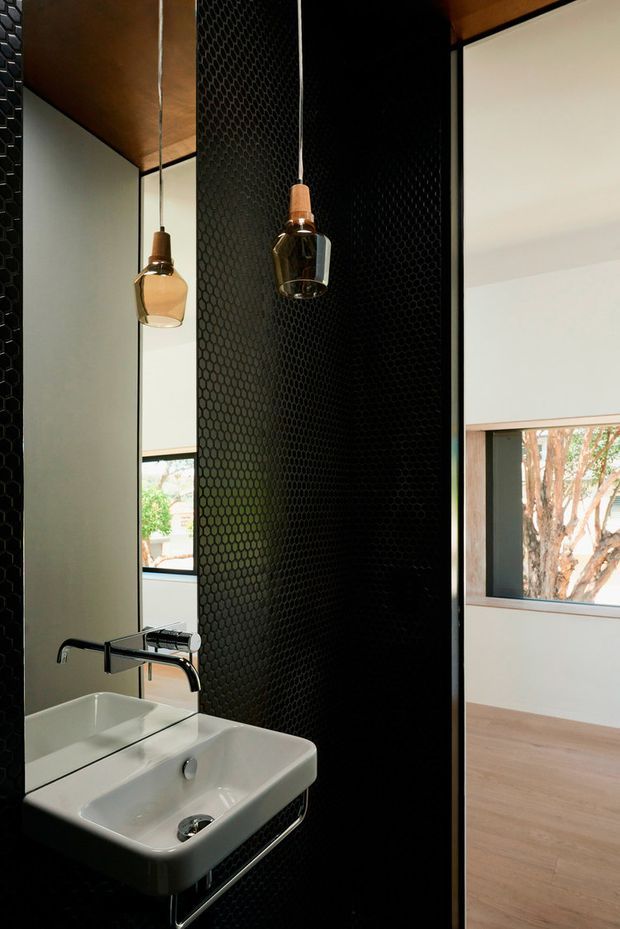 A compact contemporary bathroom featuring a wall-mounted white basin with black metal support, black wall-mounted tapware, a full-height black hexagonal textured tile feature wall, and a large mirror. Warm timber ceiling and flooring are visible, with a suspended pendant light and an opening toward an adjacent bright room.