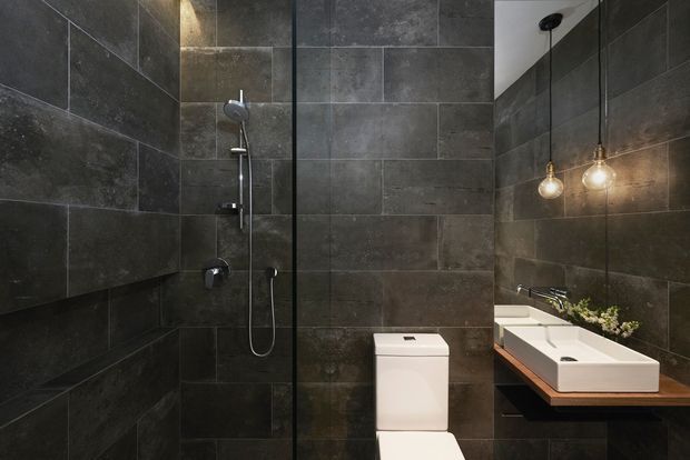 A compact contemporary bathroom with dark charcoal-grey large-format wall tiles, a glass-enclosed shower, wall-mounted shower fittings, a white toilet, and a white vessel basin set on a timber vanity shelf. Two exposed pendant bulbs provide warm accent lighting above the basin.