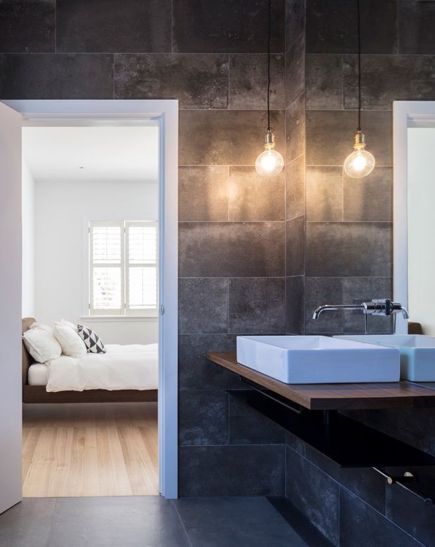 A contemporary bathroom with dark, textured large-format wall and floor tiles, a timber vanity benchtop, white vessel basins, chrome wall-mounted tapware, and twin exposed pendant lights. An open doorway reveals an adjacent bedroom with white bedding and pale timber flooring.