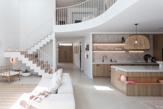Bright, open-plan contemporary interior featuring a white sculptural staircase with curved upper balustrade, a minimalist living area, and a warm timber kitchen with island bench. The kitchen includes pale stone surfaces, integrated shelving, a tiled backsplash, and a large woven pendant light. A hallway and entry are visible beyond the living space.