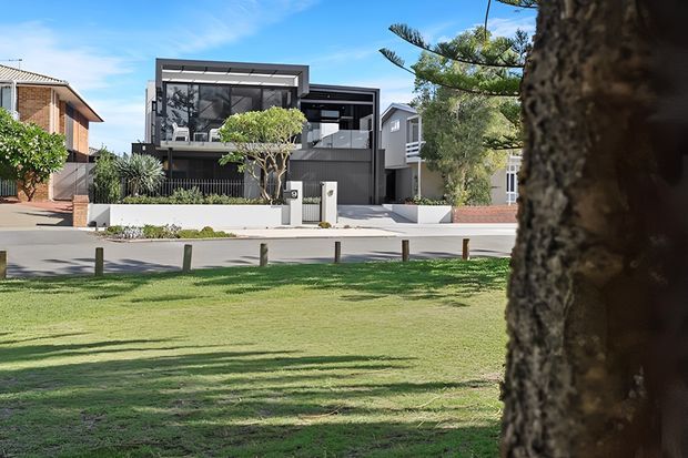Exterior street view of a contemporary multi-level dark-clad residence with a prominent upper-floor glazed terrace or balcony, flat roof forms, landscaped front garden, and a white boundary wall. The home is viewed across a grassy park-like verge with mature trees and neighboring suburban houses visible around it.