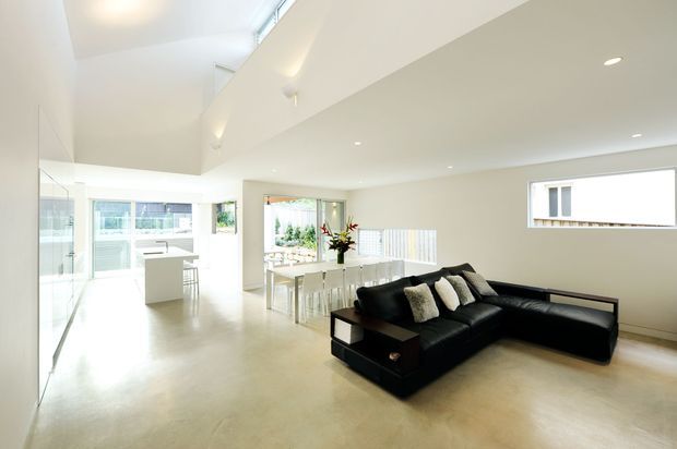 Bright, open-plan contemporary interior with a polished pale floor, white walls and ceiling, and extensive glazing. A black L-shaped sectional sofa defines the living area, while a white dining table and chairs sit beside a compact white kitchen with an island or peninsula. High-level glazing and a raised ceiling bring in abundant natural light.