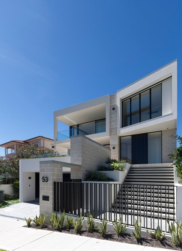 Street-level exterior view of a contemporary multi-level residence with a pale rendered and blockwork facade, strong rectilinear forms, large black-framed glazed openings, recessed balconies with glass balustrades, and a broad stepped entry behind a black metal fence. Low-maintenance landscaped planting and neighbouring homes are visible.