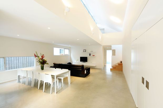 Bright open-plan modern interior combining a dining area and living room, with a white dining table and chairs in the foreground, a black sectional sofa and wall-mounted television beyond, and a compact timber staircase leading upward. The space has polished concrete flooring, white walls and built-in cabinetry, recessed lighting, and a high ceiling with a linear skylight or clerestory element. A balcony or stair void railing is visible at left.