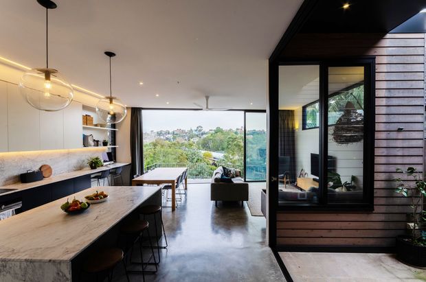 A contemporary open-plan kitchen and living area with a large stone island, dark bar stools, pendant globe lights, integrated white cabinetry, and polished concrete flooring. Full-height glazing opens to a leafy suburban outlook, while a dark timber-clad internal volume with black-framed windows creates a strong architectural feature. The space is warm, refined, and connected to the outdoors.
