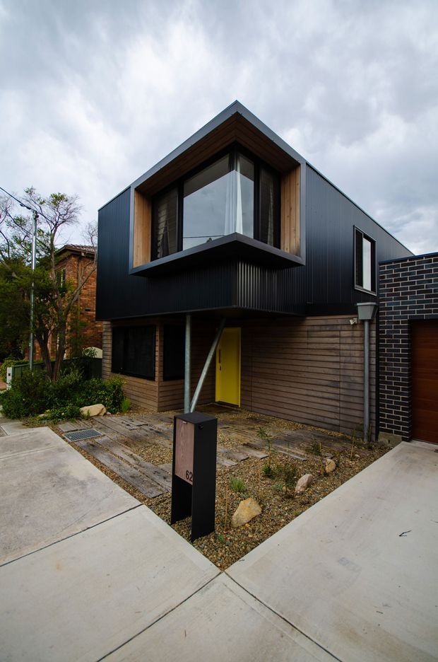 Exterior street-level view of a contemporary two-storey urban townhouse or compact residence. The building features a projecting upper-level glazed bay, dark vertically ribbed metal cladding, warm timber lining and horizontal timber siding, charcoal brickwork, and a bright yellow entry door. Concrete paths, gravel landscaping, a front mailbox, and an attached garage are visible under an overcast sky.