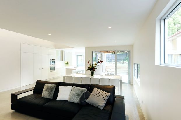 Bright open-plan interior with a black sectional sofa in the foreground, a white minimalist kitchen with integrated cabinetry and appliances, and a dining area beyond. Large glazed doors and windows provide abundant natural light and connect to an outdoor deck or terrace. The space has a clean, contemporary fit-out with white walls, pale timber flooring, recessed downlights, and restrained furnishings.