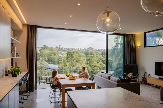 A contemporary open-plan living and dining area with a large dining table, kitchen island and bar seating, built-in shelving, and a sectional sofa. Full-height glazing overlooks a leafy suburban landscape, while sculptural glass pendant lights, recessed ceiling lighting, dark curtains, and warm timber-toned joinery create a refined, welcoming interior.