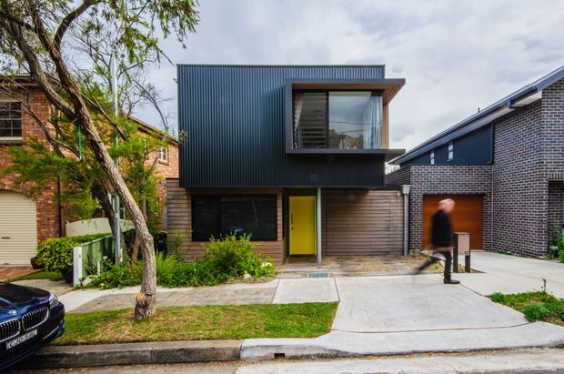 Street-facing view of a compact contemporary two-storey townhouse or residential extension. The facade combines dark vertically ribbed metal cladding on the upper level with horizontal timber-look cladding below, a projecting upper-floor window with a deep timber-toned reveal, and a vivid yellow front door. Concrete paths, a small lawn, established street tree, neighbouring brick homes, and a blurred pedestrian are visible.