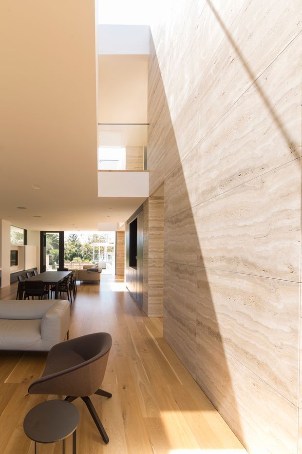 A bright, double-height open-plan living and dining area with warm timber flooring, a large horizontally layered natural stone feature wall, and minimalist built-in joinery. A dining table and chairs sit beside a pale sofa and sculptural lounge chair, while full-height glazing opens toward a leafy outdoor setting. A mezzanine or upper-level opening is visible above the living area, with strong natural light creating dramatic shadows across the stone wall.