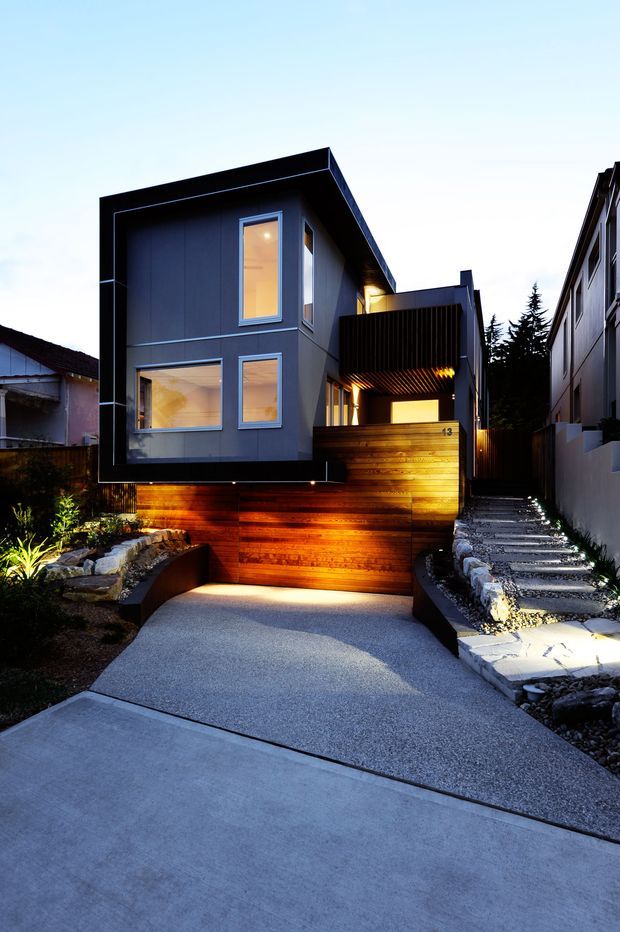 Twilight exterior view of a compact contemporary multi-level residence on a narrow urban site. The dark, sharply articulated box-like facade features large rectangular windows, a projecting flat roof, and a warm timber-clad lower level and entry screen. A textured driveway leads to the house, with a raised illuminated timber wall, landscaped rock beds, and a narrow stepped side walkway.