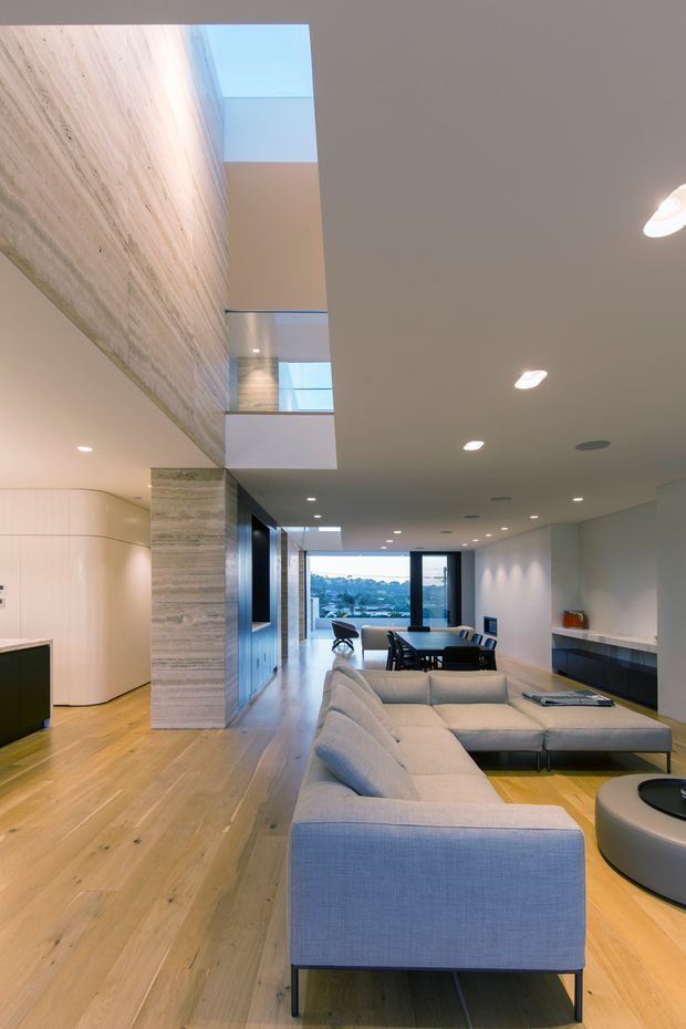 A refined open-plan contemporary interior viewed from the living area toward a dining space and expansive glazed opening. The room features a large pale upholstered sectional sofa, light timber flooring, a long dark built-in media/storage unit, recessed ceiling lights, and a dramatic double-height void with a large skylight. A textured pale stone or stone-look wall rises through the void, while curved pale cabinetry is visible to the left. The space is bright, spacious, and minimalist, with an urban outlook beyond the glazing.