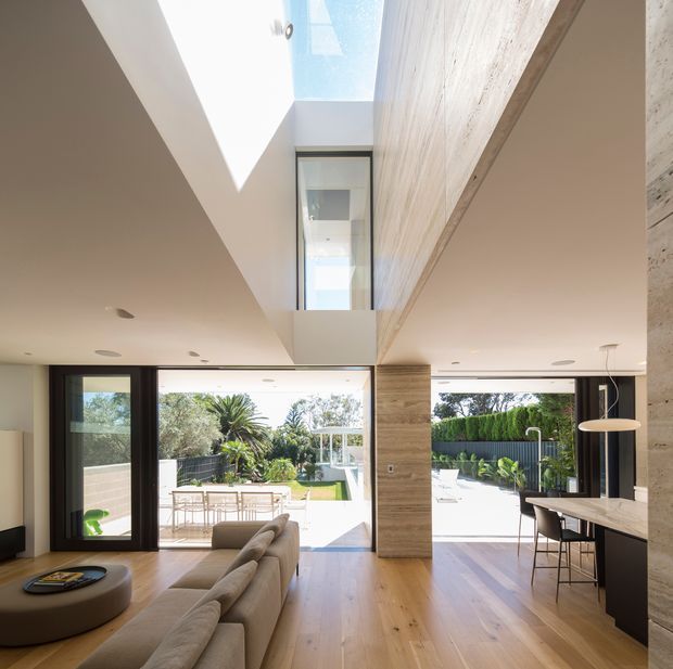 Bright contemporary open-plan living and dining space with warm timber flooring, expansive black-framed glazing opening to a landscaped backyard and pool area, a double-height void with skylight, and a large textured stone feature wall. A sofa and coffee table occupy the living zone, while a dining or breakfast bar with stools sits to the right.