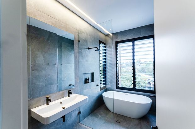Contemporary bathroom with a freestanding white bathtub beneath a large black-framed louvred window, a wall-mounted rectangular vanity, frameless glass shower enclosure, dark tapware, and grey stone-look wall and floor finishes. Integrated linear lighting and clean geometric detailing create a refined, modern interior.