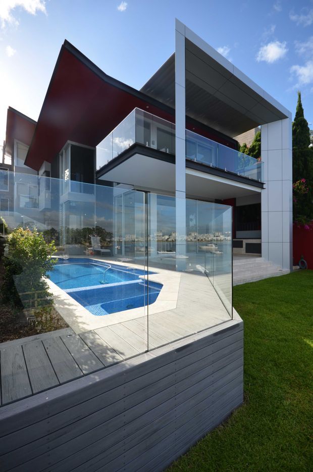 Exterior view of a contemporary multi-level residence beside a swimming pool, featuring dramatic angular roof forms, bold red and dark facade surfaces, extensive floor-to-ceiling glazing, glass balustrades, and a large cantilevered roof canopy. The landscaped outdoor area includes a timber-clad raised deck and transparent pool fencing, with a waterfront or urban skyline visible through the glazing.