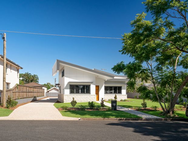 Street-facing view of a contemporary single-storey house with a white rendered facade, low-pitched skillion-style roof forms, broad eaves, and light stone-clad lower walls. The property includes a paved driveway, front lawn, landscaped garden beds, and a small detached garage or outbuilding visible to the rear. Clear blue sky and mature street trees surround the home.
