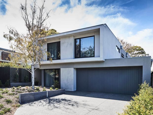 A contemporary two-storey residence viewed from the street, with a crisp white rendered facade, dark-framed windows, a large charcoal double garage, and a broad concrete driveway. The house has a simple gabled roof form, projecting upper-level volume, landscaped front garden beds, and a dark boundary fence.
