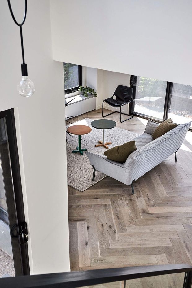 A bright contemporary living room viewed from an adjacent stair or landing, featuring a white textured sofa with olive cushions, two small round coffee tables, a black lounge chair, a pale area rug, and herringbone timber flooring. Large black-framed windows and sliding glazing bring in abundant natural light, while a simple pendant bulb hangs in the foreground.