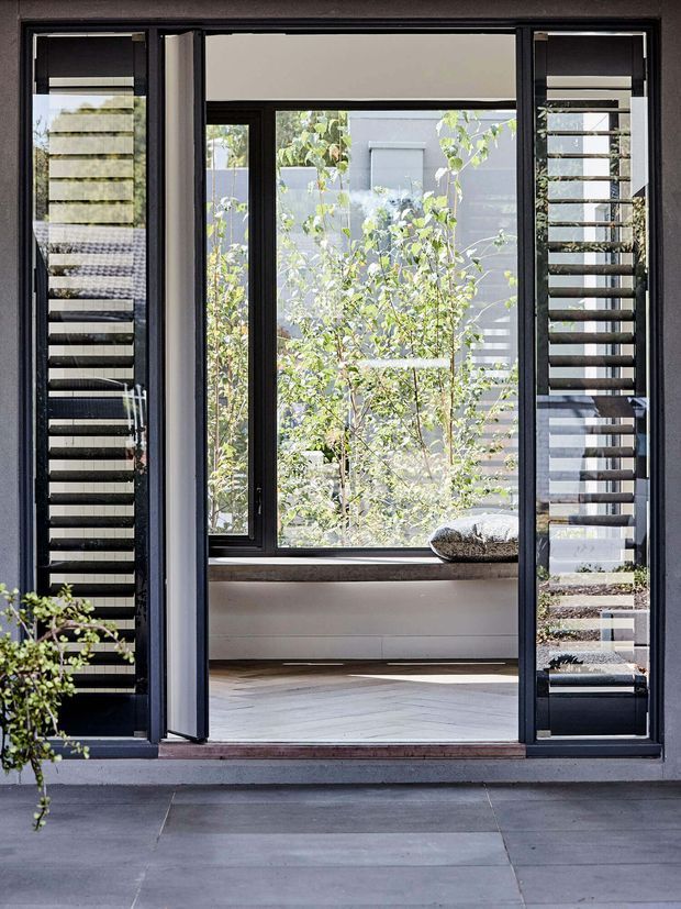 A modern black-framed glazed doorway viewed from outside, with open central doors and fixed louvred side panels. The entry leads to a bright enclosed sitting area with a built-in window seat, pale timber herringbone flooring and abundant garden greenery beyond.