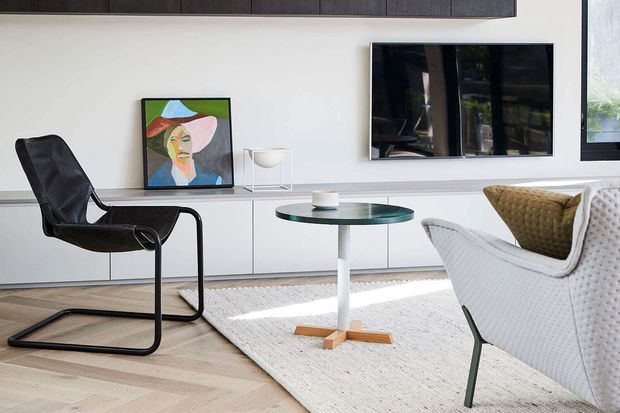 A bright, contemporary living room with a pale timber floor, white built-in media cabinetry, wall-mounted television, and large windows. A black sling lounge chair, white textured armchair, round glass coffee table, woven rug, and colourful artwork create a clean modern interior with contrasting dark accents.