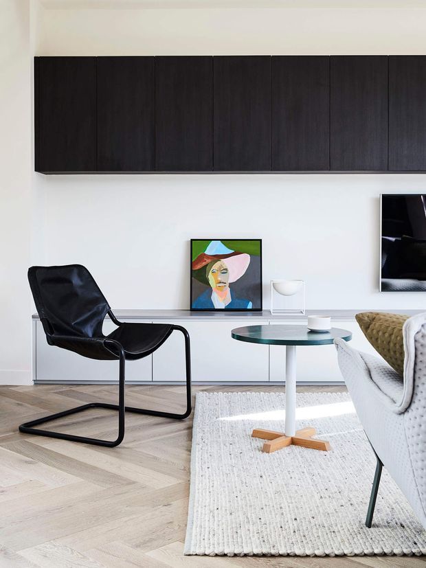 Bright, refined contemporary living room with pale herringbone timber flooring, white walls, extensive dark timber wall cabinetry, a low white built-in media console, black leather lounge chair, glass-topped coffee table, and a light upholstered sofa. The room has a clean, curated aesthetic with artwork and restrained furnishings.