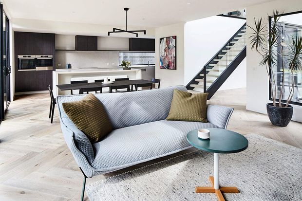 Bright open-plan contemporary interior combining a living room, dining area and kitchen. A pale textured sofa and round coffee table sit on a large rug in the foreground, with a dark timber kitchen, white tiled splashback, dining table, pendant lighting and an open staircase beyond. Large glazing and indoor plants add an airy feel.