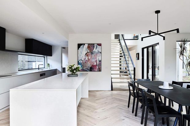 Bright open-plan contemporary kitchen and dining area with a large white island, dark integrated cabinetry, pale herringbone timber flooring, black dining furniture, and a sculptural black linear pendant light. A staircase with open risers and black detailing leads upward, while large windows and glazed doors bring in natural light.