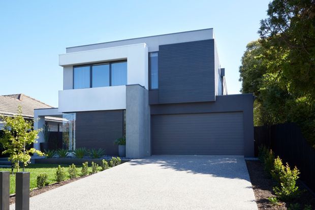 Contemporary two-storey suburban house with a crisp geometric facade, white rendered upper volumes, dark horizontal cladding, concrete-look vertical feature wall, large glazed openings, and an integrated double garage. A broad light gravel driveway leads to the entrance, with simple landscaped garden beds and mature trees surrounding the property.
