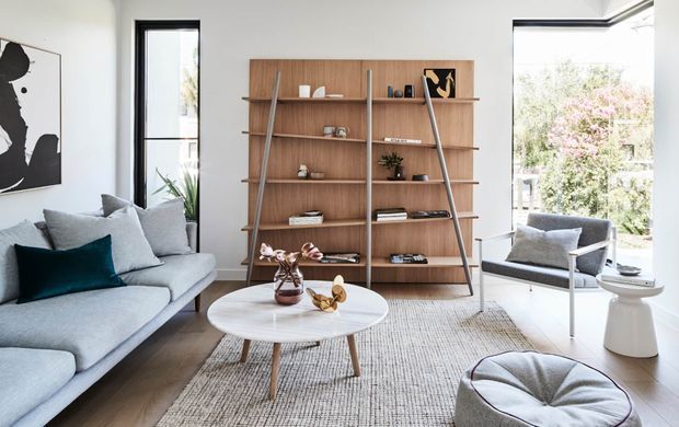 Bright, refined living room with a large light-grey sectional sofa, sculptural round coffee table, textured rug, and a prominent built-in timber shelving unit. Floor-to-ceiling corner glazing brings in abundant natural light and views of greenery, while contemporary artwork, cushions, and restrained decorative objects add subtle character.