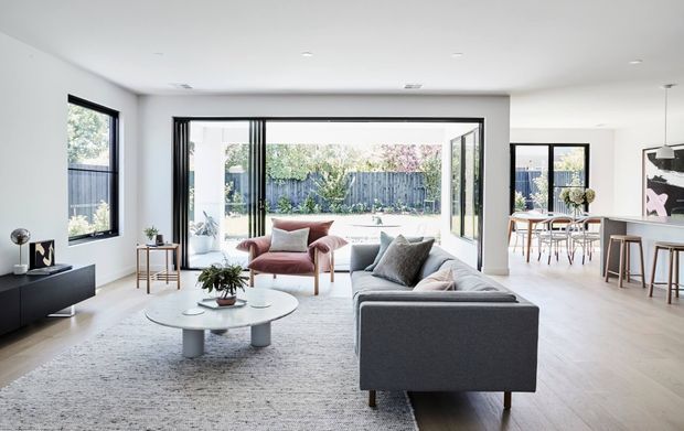 Bright open-plan contemporary living space with a grey sectional sofa, blush lounge chairs, round coffee table, pale timber flooring, and a large textured rug. Full-width sliding glass doors open the living room to a landscaped backyard, while a dining area and kitchen island are visible to the right. The interior has white walls, minimal detailing, and abundant natural light.