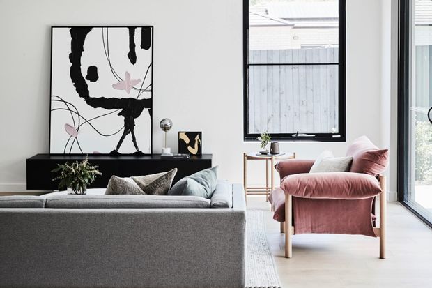A bright contemporary living room viewed from behind a large grey sofa. The space features a black low-profile media console, oversized abstract artwork, a blush pink upholstered lounge chair, pale timber side table, neutral rug, indoor plant and extensive black-framed glazing. The palette is soft and refined with strong black-and-white graphic accents.