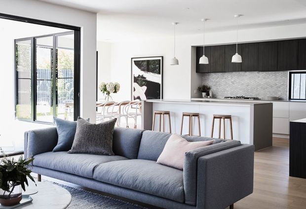 Bright open-plan contemporary interior featuring a large blue-grey sectional sofa in the foreground, a white kitchen island with timber stools, dark timber cabinetry, a light stone-textured splashback, pendant lights, and expansive black-framed glazing opening toward an outdoor area.