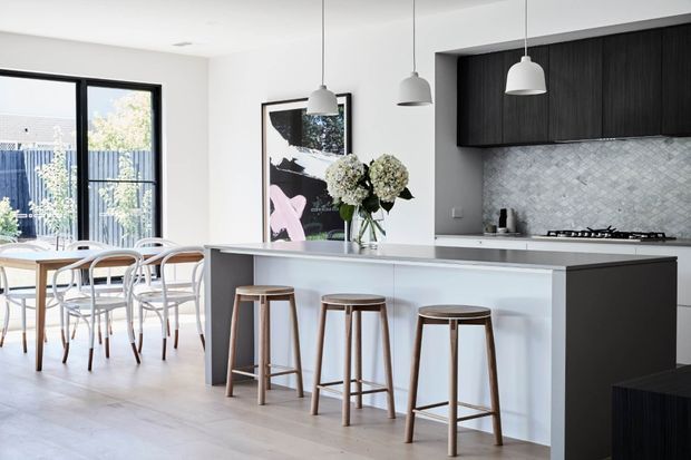 Bright contemporary open-plan kitchen and dining area with a long white island, integrated dark timber cabinetry, pale textured stone-look splashback, and three suspended grey pendant lights. The island has three timber stools, while a nearby dining table sits beside large black-framed glazing. A floral arrangement and artwork add decorative accents.