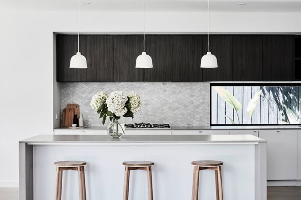 Bright contemporary kitchen featuring a large white island with three timber stools, dark vertical-grain overhead cabinetry, a pale patterned tile splashback, integrated cooktop, and three white pendant lights. A horizontal window brings in natural light and views of foliage.