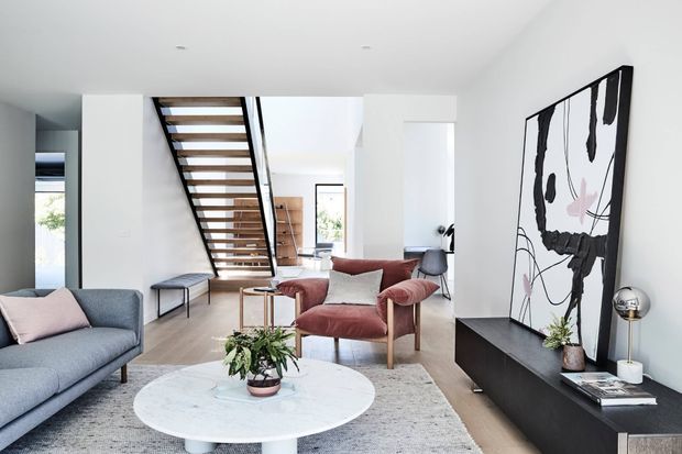 Bright contemporary open-plan living room with a grey sofa, blush upholstered armchairs, a round white coffee table, pale timber flooring, and a large abstract artwork above a low black media console. An open-riser timber staircase with dark metal framing leads to the upper level, and white walls and recessed lighting create a clean, airy setting.