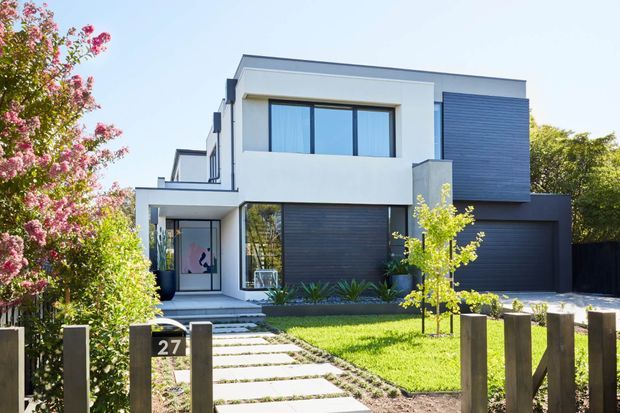 Street-facing view of a contemporary two-storey residence with a white rendered facade, dark horizontal cladding, large upper-level glazing, an integrated garage, and a landscaped front garden with a paved entry path and timber fence.