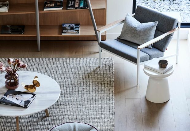 A refined contemporary living room corner featuring a grey upholstered armchair with light timber arms, a round white coffee table, a textured neutral rug, pale timber flooring, and built-in timber shelving with books and decor. A small white side table and large window complete the bright, understated setting.