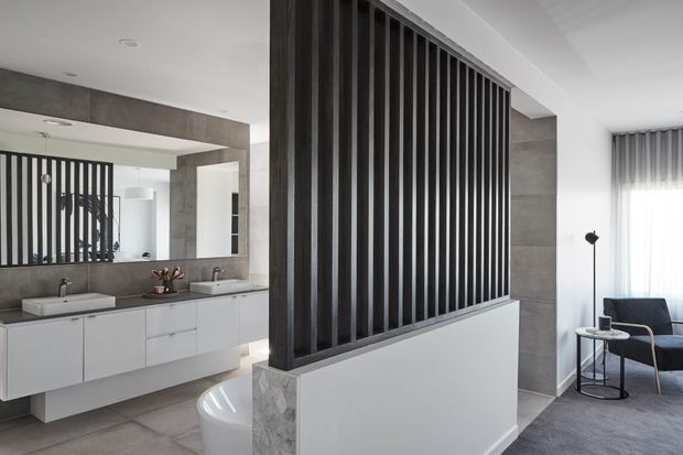 Contemporary open-plan bathroom with a floating white vanity, twin basins, large wall mirror, freestanding bathtub and a striking dark vertical-slat privacy screen. A small adjoining lounge area is visible beyond the partition, with grey stone-look wall finishes, grey flooring and soft natural light.