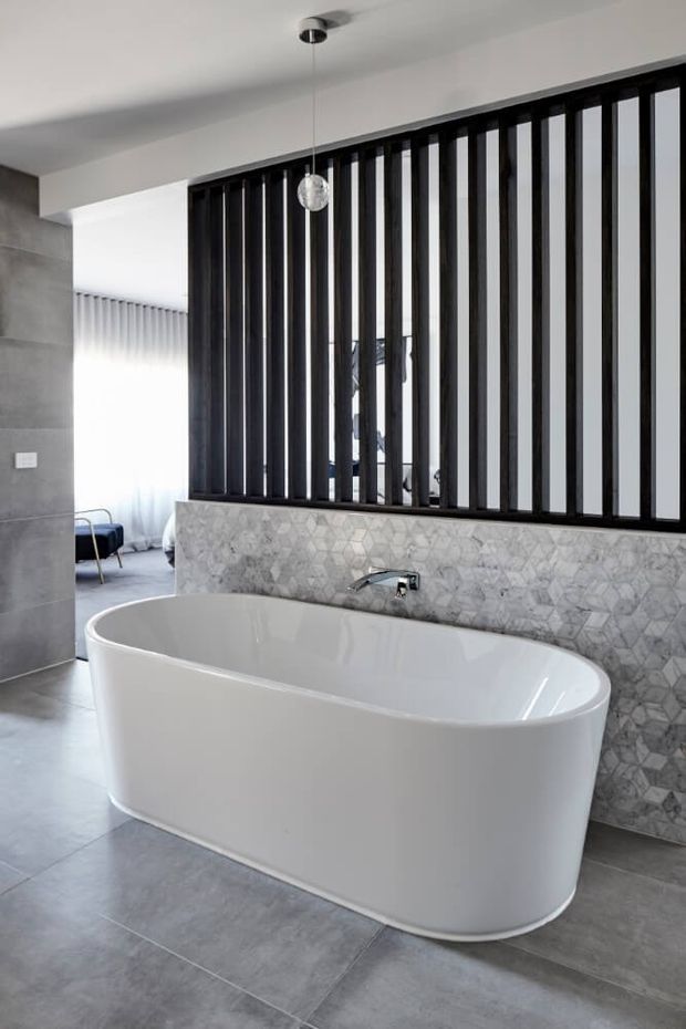 Contemporary bathroom featuring a large freestanding white bathtub positioned in front of a grey geometric stone-tile feature wall. A black vertical-slat screen separates the bath from an adjoining bedroom or living area, with grey tiled flooring, a wall-mounted chrome tap, and a minimalist pendant light.