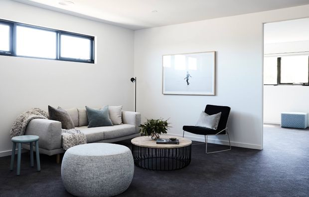 A bright, minimalist living room with a pale grey sofa, textured round ottoman, black wire coffee table, black lounge chair, and a small side table. White walls, high horizontal windows, framed artwork, and grey carpet create a calm contemporary interior. An adjoining carpeted room is visible through the wide opening.