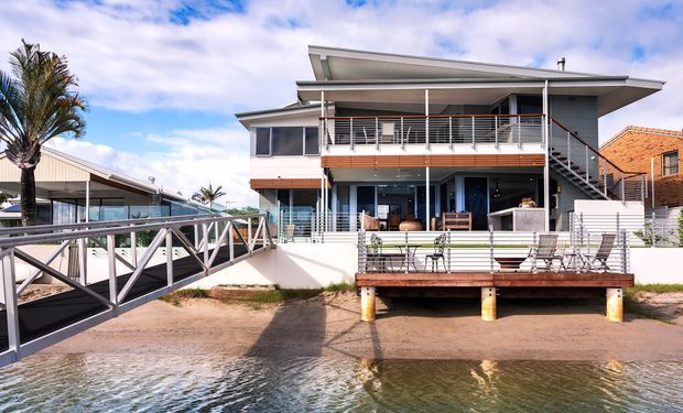Exterior view of a contemporary multi-level waterfront residence with broad roof overhangs, extensive balconies and decks, metal balustrades, glazed openings, and a raised structure positioned directly beside the water. A connecting pedestrian bridge and neighboring buildings are also visible.