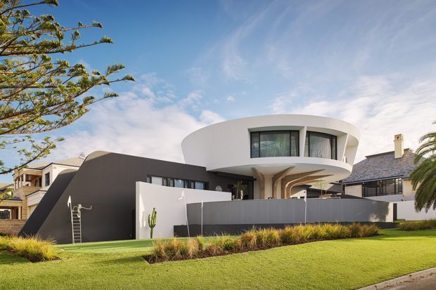 Exterior view of a striking contemporary multi-level residence with a rounded white upper volume projecting over a dark charcoal lower wing. The house features curved forms, broad glazing, a flat roof, and a restrained landscaped front lawn with ornamental grasses. Traditional neighbouring homes are visible on either side.