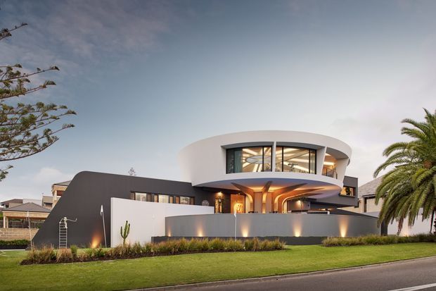 Contemporary two-storey residence with a sculptural curved upper level, broad arched glazing, dark and white rendered facades, and a sheltered entry beneath a dramatic cantilevered form. The home is presented from the street with a manicured lawn, low boundary wall, architectural planting, and palm trees.