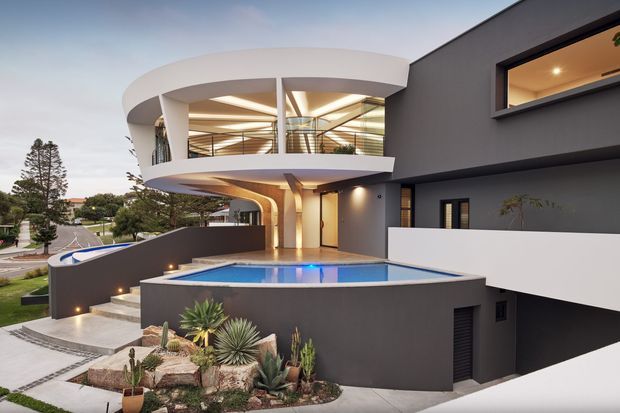 Contemporary multi-level residential home viewed from the street, featuring bold curved white balcony forms, dark rendered walls, extensive glazing, recessed lighting, and a raised rectangular swimming pool. Drought-tolerant succulent landscaping and a gently sloping suburban street surround the property.