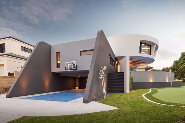 Exterior view of a contemporary multi-level residence with sculptural angular concrete or rendered walls, a curved upper-floor form, dark privacy screens, and expansive glazing. A swimming pool, paved terrace, manicured lawn, and basketball hoop occupy the foreground, with warm interior lighting visible beneath the curved cantilevered section.